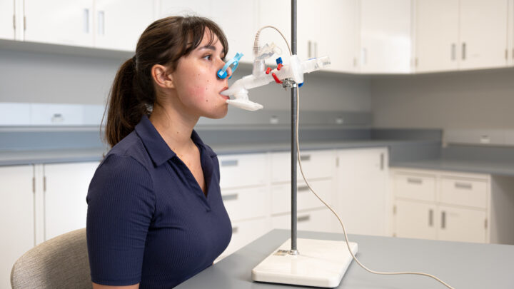 A woman sits at a table in a clinical room. On the table is a medical apparatus on a stand, with a tube extended from it that she is holding in her mouth. She has a light blue clamp over her nose.